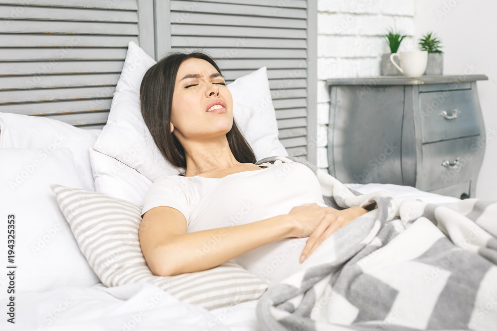 Young woman suffering from abdominal pain while sitting on bed at home. Stomach pain. In stress. Young woman suffering from abdominal pain while sitting on bed at home. Stomach pain. In stress.