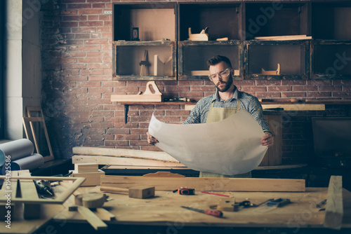 Front view portrait of thoughtful minded serious pensive confident concentrated furniture maker wearing checkered casual shirt apron and protective glasses, he is holding a blueprint with instruction