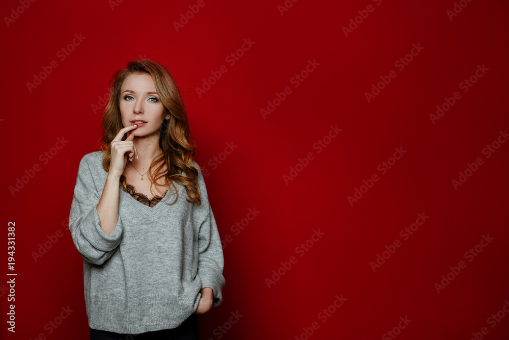 Body language and emotions.Portrait of beautiful cheerful blonde girl with curly hair over red background.The woman holding her finger on teeth making difficult choice not knowing what to choose. Body language and emotions.Portrait of beautiful cheerful blonde girl with curly hair over red background.The woman holding her finger on teeth making difficult choice not knowing what to choose.