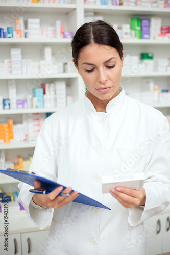 Beautiful young pharmacist holding clipboard and a box of medications