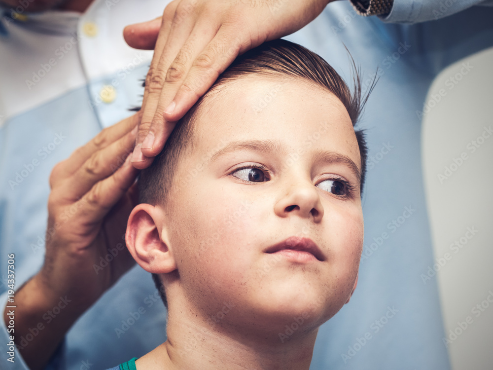 Naklejka premium Barber is making a hairstyle to Caucasian boy in barbershop.