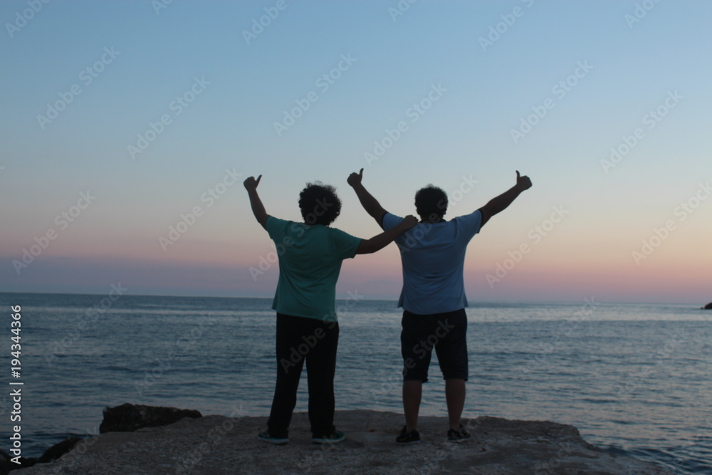 Fototapeta premium two young men thumb up near sea, background is sunset