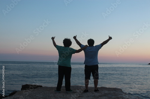 two young men thumb up near sea, background is sunset