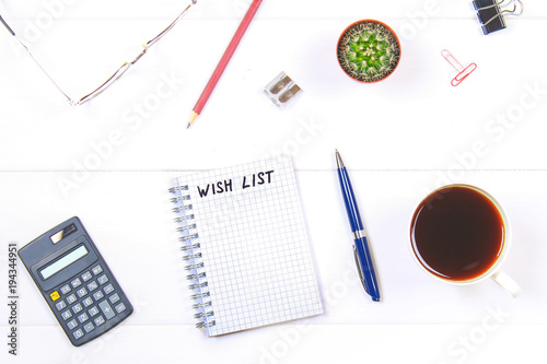 Notepad with text: wish list. White table with calculator, cactus, note paper, coffee mug, pen, glasses.