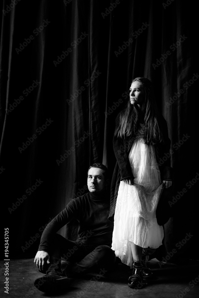 Beautiful couple on a black background. A girl in a white dress is standing and looking out the window, and the guy is sitting on the floor next to her. B&W Beautiful couple on a black background. A girl in a white dress is standing and looking out the window, and the guy is sitting on the floor next to her. B&W