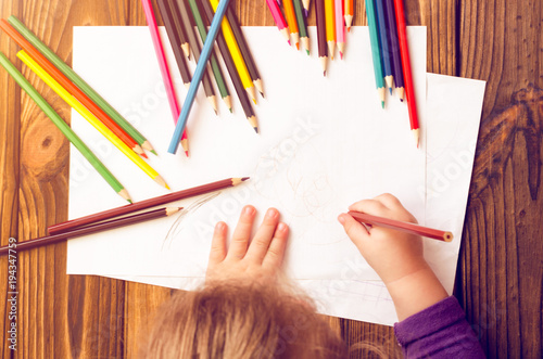 The child's hands are painted with colored pencils on a white sheet of paper on a wooden table