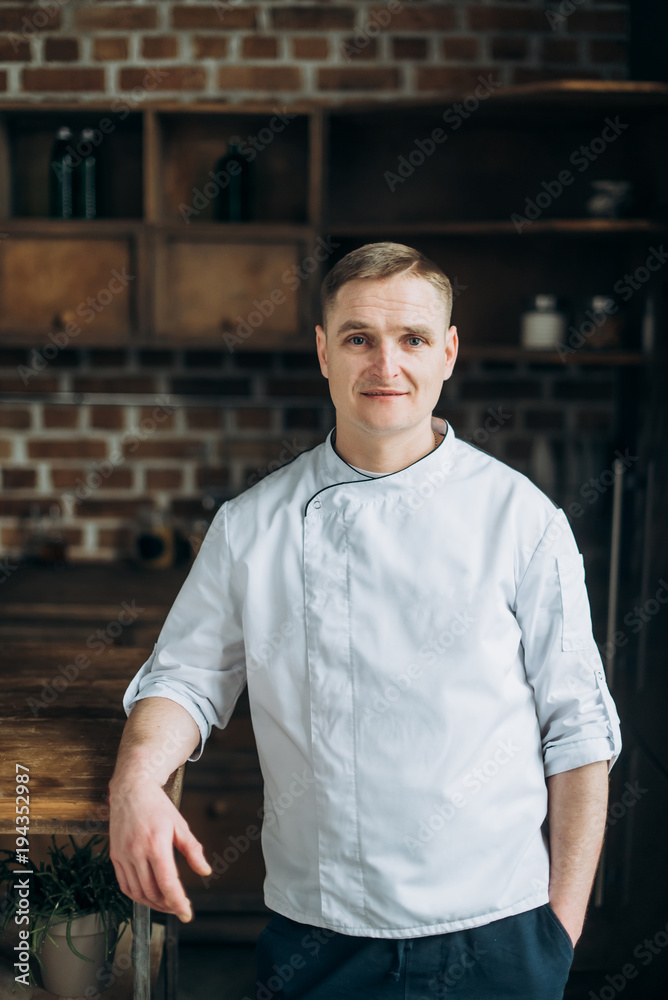 Fototapeta premium Portret charyzmatycznego kucharza o niebieskich oczach, który stoi w kuchni w białym mundurze