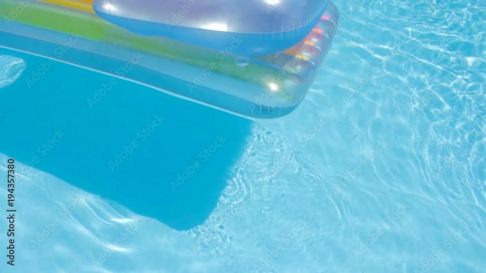 Pool raft serenely floats past the camera on the surface of a swimming pool at a water park. The crystal clear water sparkles and refracts in the bright sunlight. The shot embodies relaxation and para
