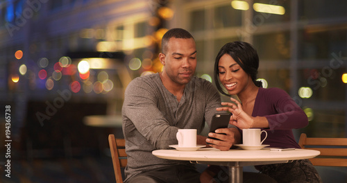 Happy Black couple using smart phone in coffee shop at night. African American man and woman using cellphone while drinking in cafe during evening. Millennials dating