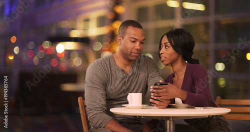 Happy Black couple using smart phone in coffee shop at night. African American man and woman using cellphone while drinking in cafe during evening. Millennials dating