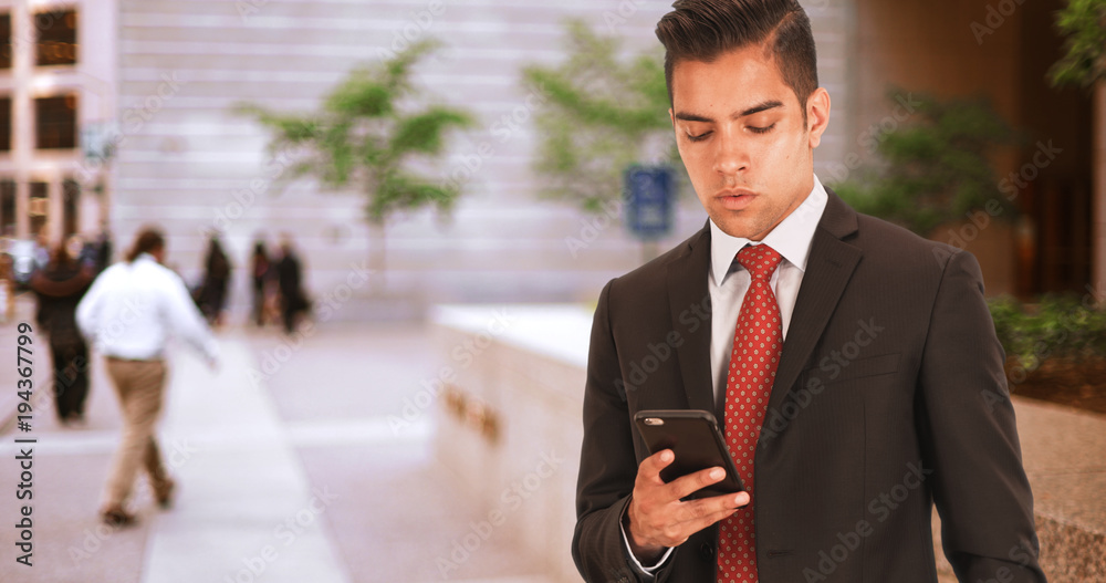 Businessman sms texting or using social media on cell phone in front of office building. Hispanic millennial man outside in suit and tie with copy space and smartphone Businessman sms texting or using social media on cell phone in front of office building. Hispanic millennial man outside in suit and tie with copy space and smartphone
