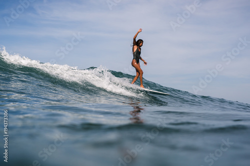 Beautiful young indonesian woman in bikini surfing wave in Bali on the background of blue sky, clouds and tropical beach