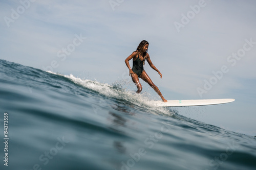 Beautiful young indonesian woman in bikini surfing wave in Bali on the background of blue sky, clouds and tropical beach