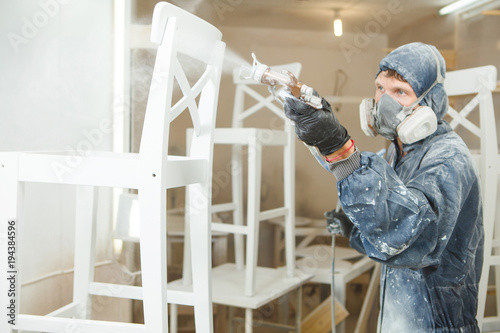 Man painting chair into white paint in respiratory mask. Application of flame retardant ensuring fire protection, airless spraying. photo