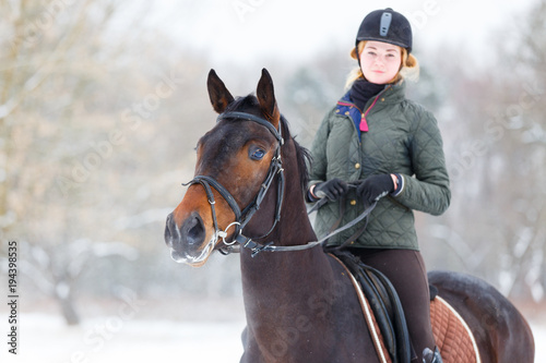 Young happy woman riding her bay horse on winter field. Winter equestrian activity background