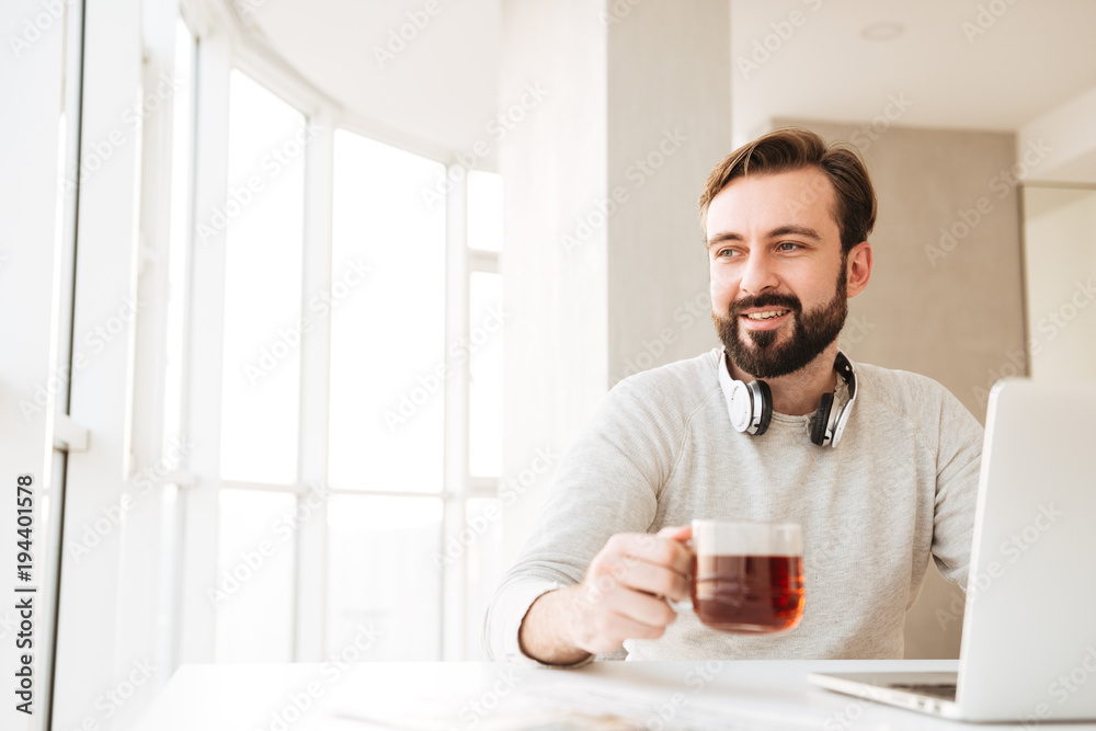 Attractive man with short brown hair and beard, drinking tea and looking through window while working on notebook in modern office