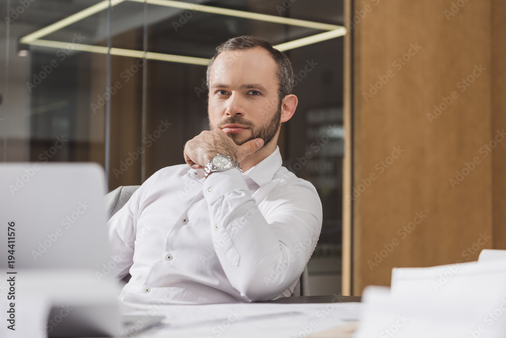 Naklejka premium handsome thoughtful architect sitting at workplace and looking at camera