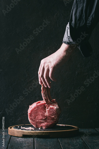 Man's hands holding raw uncooked black angus beef tomahawk steak on bone with salt and pepper on round wooden slate cutting board over dark wooden plank table. Rustic style. Toned image