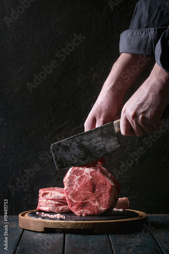 Man's hands cutting raw uncooked black angus beef tomahawk steaks on bones by vintage butcher cleaver on round wooden slate cutting board over dark wooden plank table. Rustic style