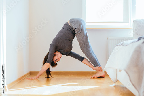 Young attractive smiling woman practicing yoga, doing downward dog facing exercise, stretching back, working out, wearing sportswear, grey pants, indoor full length, home interior.