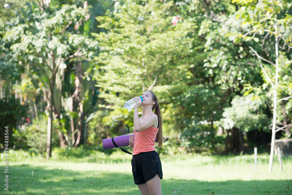 Beautiful young sportswoman is holding Yoga Mat and drink water at the park. Health and Fitness concept Beautiful young sportswoman is holding Yoga Mat and drink water at the park. Health and Fitness concept