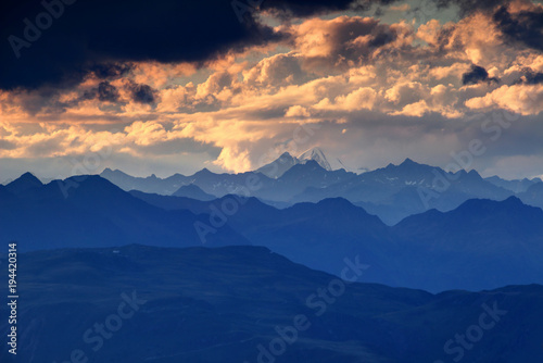 Jagged blue ridge silhouettes, snowcapped Rotspitze Pizzo Rosso peak and fluffy orange clouds at sunset, Venediger Group and Villgraten Mountains Defereggen Alps Hohe Tauern Osttirol Austria Europe