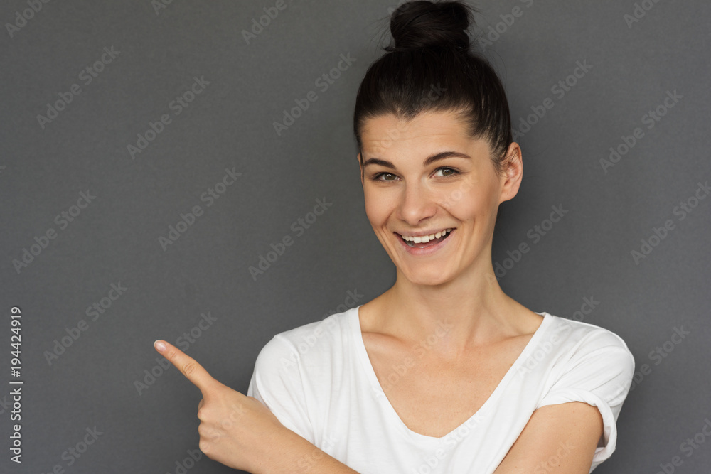 Happy young brunette Caucasian female wears white T-shirt, showing something interesting at copy space on gray wall for your information, advertising content, looking at the camera, pointing sideways. Happy young brunette Caucasian female wears white T-shirt, showing something interesting at copy space on gray wall for your information, advertising content, looking at the camera, pointing sideways.