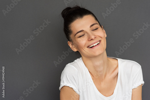 Portrait of cheerful and charming young Caucasian woman with hair bun wearing stylish white T-shirt, smiling, narrowing her eyes, looking happy. Cute positive brunette female with healthy teeth smile.