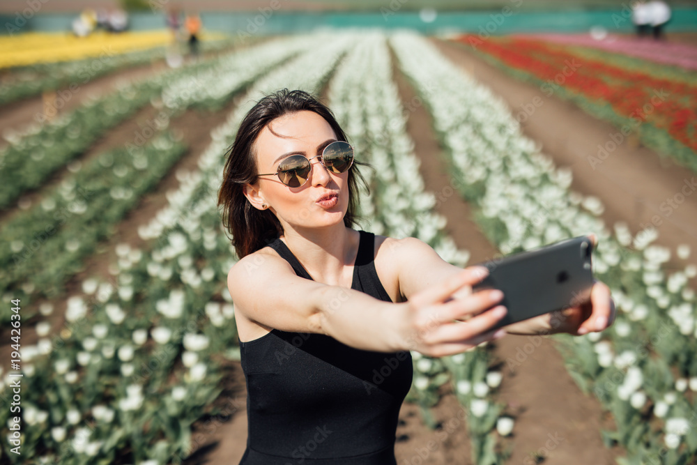Young beautiful woman wear sunglasses make selfie in tulips flower field in sunny spring day Young beautiful woman wear sunglasses make selfie in tulips flower field in sunny spring day