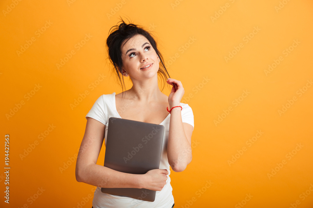 Photo of dreaming adult girl with trendy hairstyle hugging silver notebook and looking aside with lovely smile, over yellow wall copy space Photo of dreaming adult girl with trendy hairstyle hugging silver notebook and looking aside with lovely smile, over yellow wall copy space