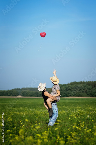 Beautiful happy young couple have fun in summer sunny field together. People wear cowboy hats. Cowgirl and cowboy with red balloon in shape of heart. Love, happiness concept. Vertical photography.