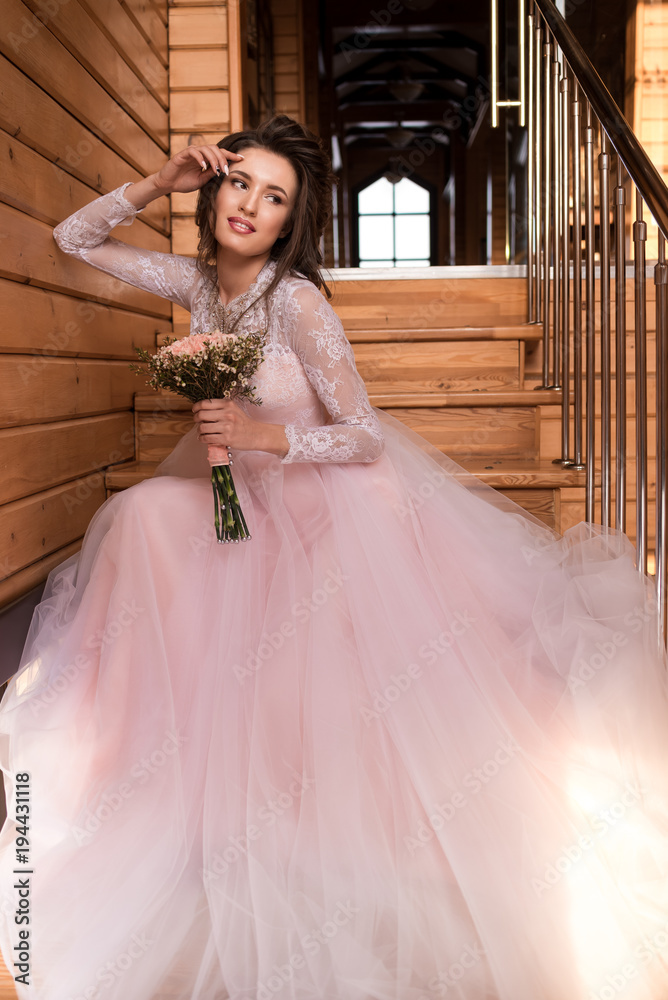Beautiful bride is sitting on the stairs with a bouquet. Hand at the face.  Wedding lace light pink dress. Wedding hairstyle and makeup. Wooden  staircase. Smiling. Stock-bilde | Adobe Stock