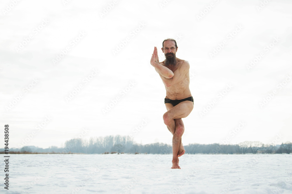 A middle-aged man is engaged in yoga on a frozen river. Vrishasana (Tree pose).