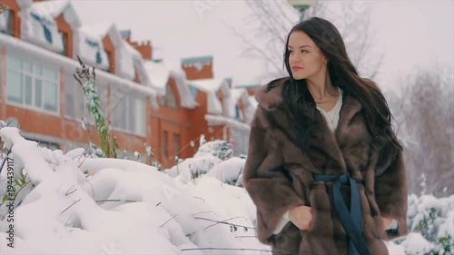Brunette Fashionable sexy pretty rich Young woman in fur mink fur coat smiling playfully looks at camera on background of beautiful buildings on a snow-covered winter street. slow motion