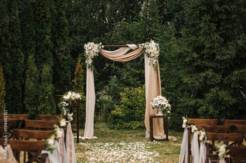 Very beautiful outside ceremony. classical wedding in forest. The arch is wooden. White flowers. Brown chairs. Rite. Bride and groom. Decor. Floristics. In the open air. Path from the petals of roses.