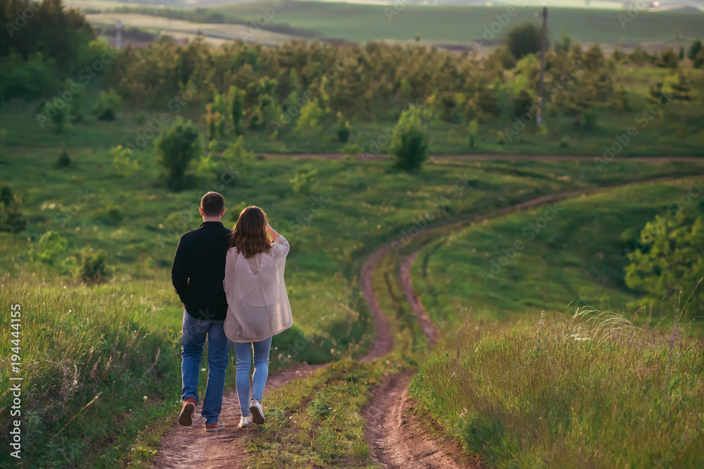 Loving boyfriend with a girlfriend walking along a road that descends from a hill in a green field, holding hands