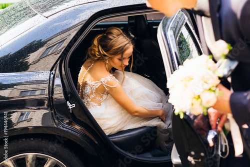 Fotografie the beautiful bride sits in a black car and the groom wants to close the door af
