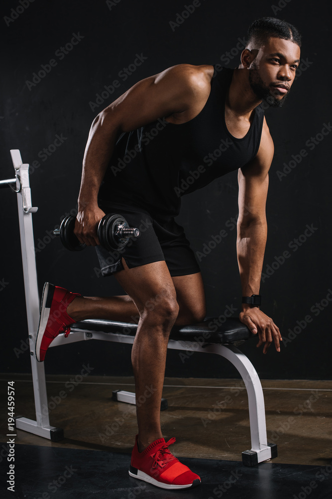 handsome muscular african american man in sportswear training with dumbbell and looking away handsome muscular african american man in sportswear training with dumbbell and looking away