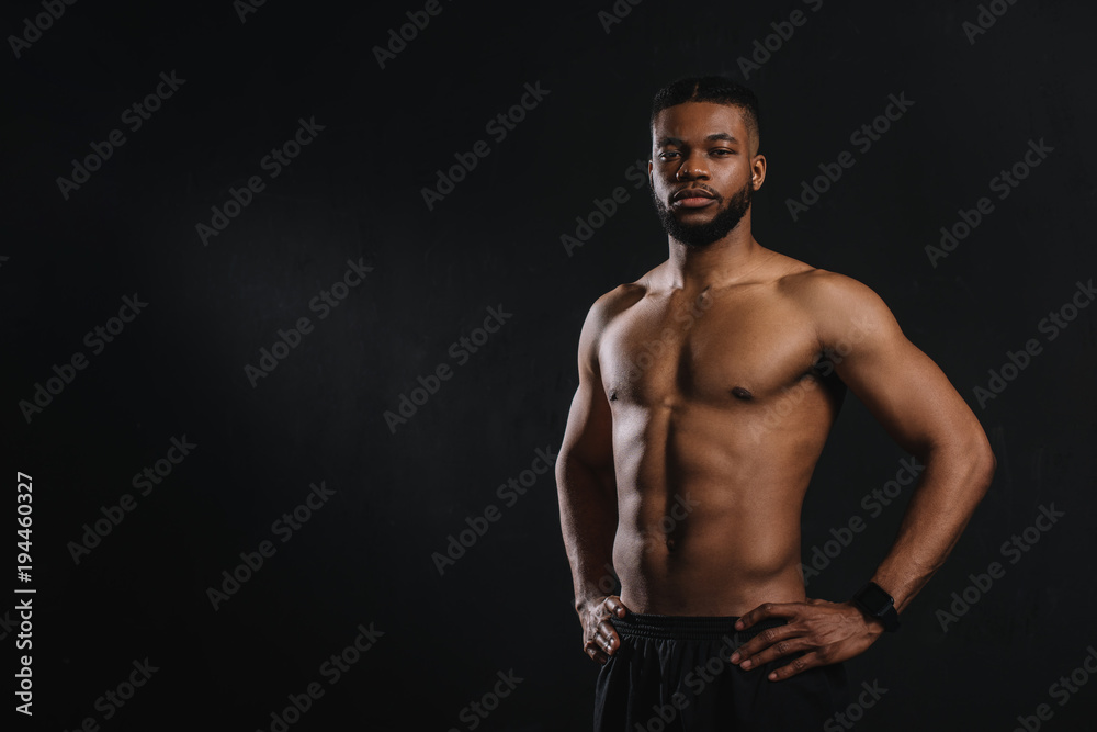 muscular shirtless african american man standing with hands on waist and looking at camera isolated on black