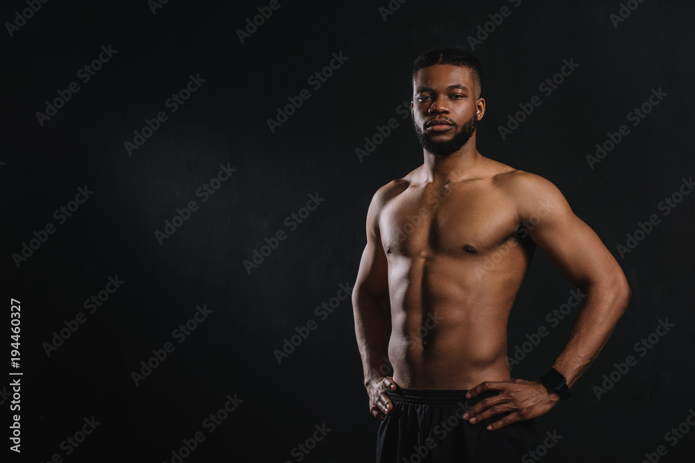 muscular shirtless african american man standing with hands on waist and looking at camera isolated on black