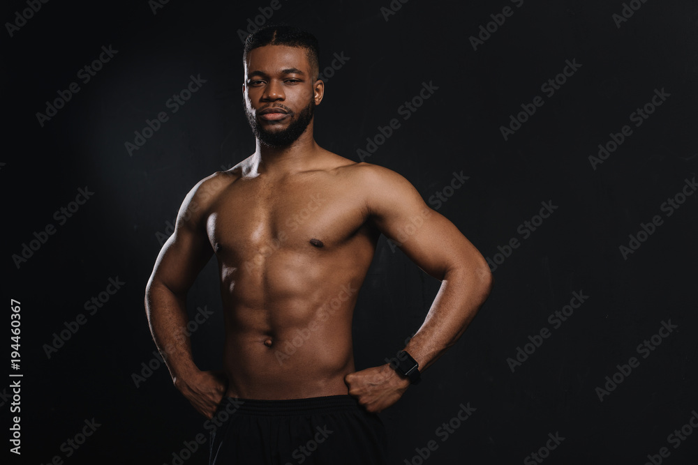 muscular young african american sportsman standing with hands on waist and looking at camera isolated on black