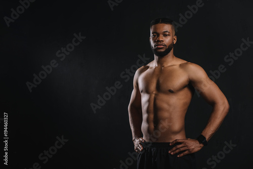 muscular shirtless african american man standing with hands on waist and looking at camera isolated on black