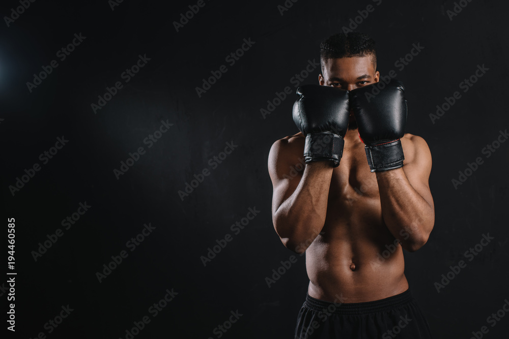 young shirtless african american boxer in boxing gloves looking at camera isolated on black