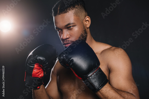 serious shirtless african american boxer in boxing gloves looking away on black