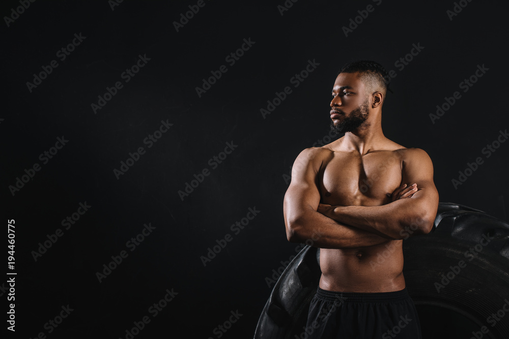 muscular shirtless african american sportsman standing with crossed arms near big tyre and looking away isolated on black