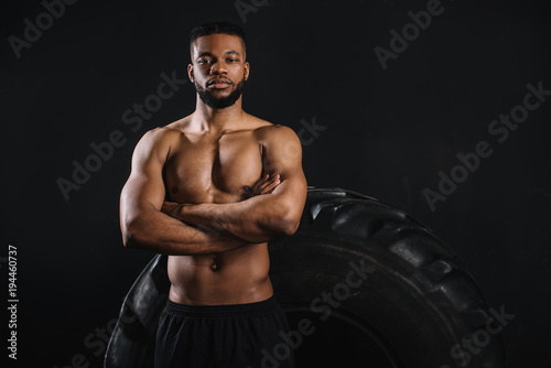 muscular shirtless african american sportsman standing with crossed arms near big tyre and looking at camera isolated on black