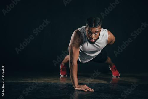 young african american sportsman doing push-ups and looking at camera on black