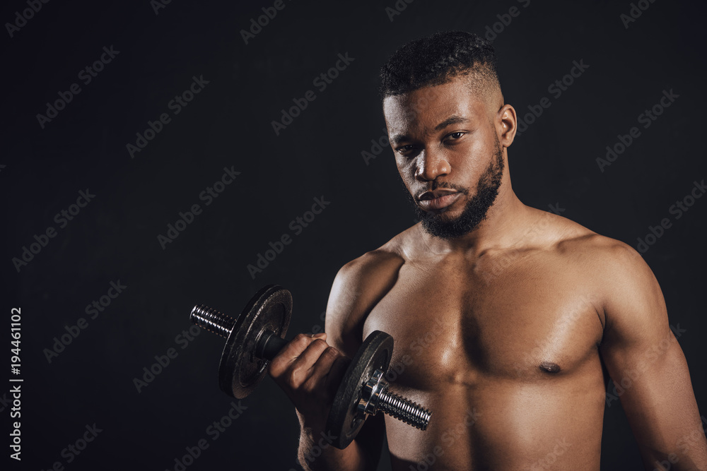 muscular shirtless african american sportsman training with dumbbell and looking at camera isolated on black muscular shirtless african american sportsman training with dumbbell and looking at camera isolated on black
