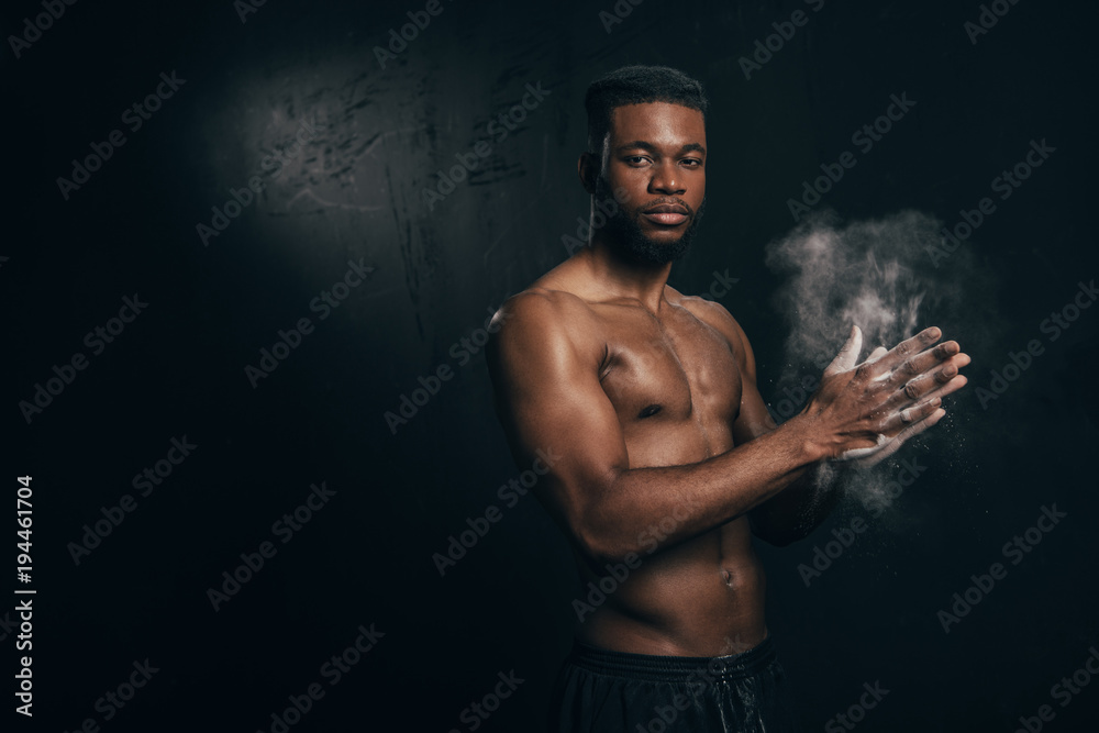 shirtless young african american sportsman applying talcum to hands and looking at camera on black
