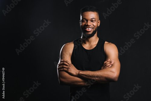 happy young african american sportsman standing with crossed arms isolated on black